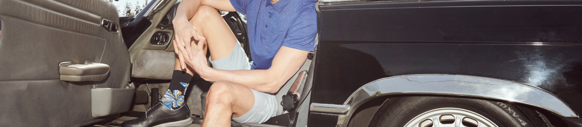 Man in short and shirt and socks sitting in driver’s side door of vehicle