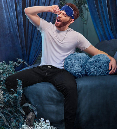A yawning man sitting on a bed while wearing a sleep mask and cooling sleepwear 