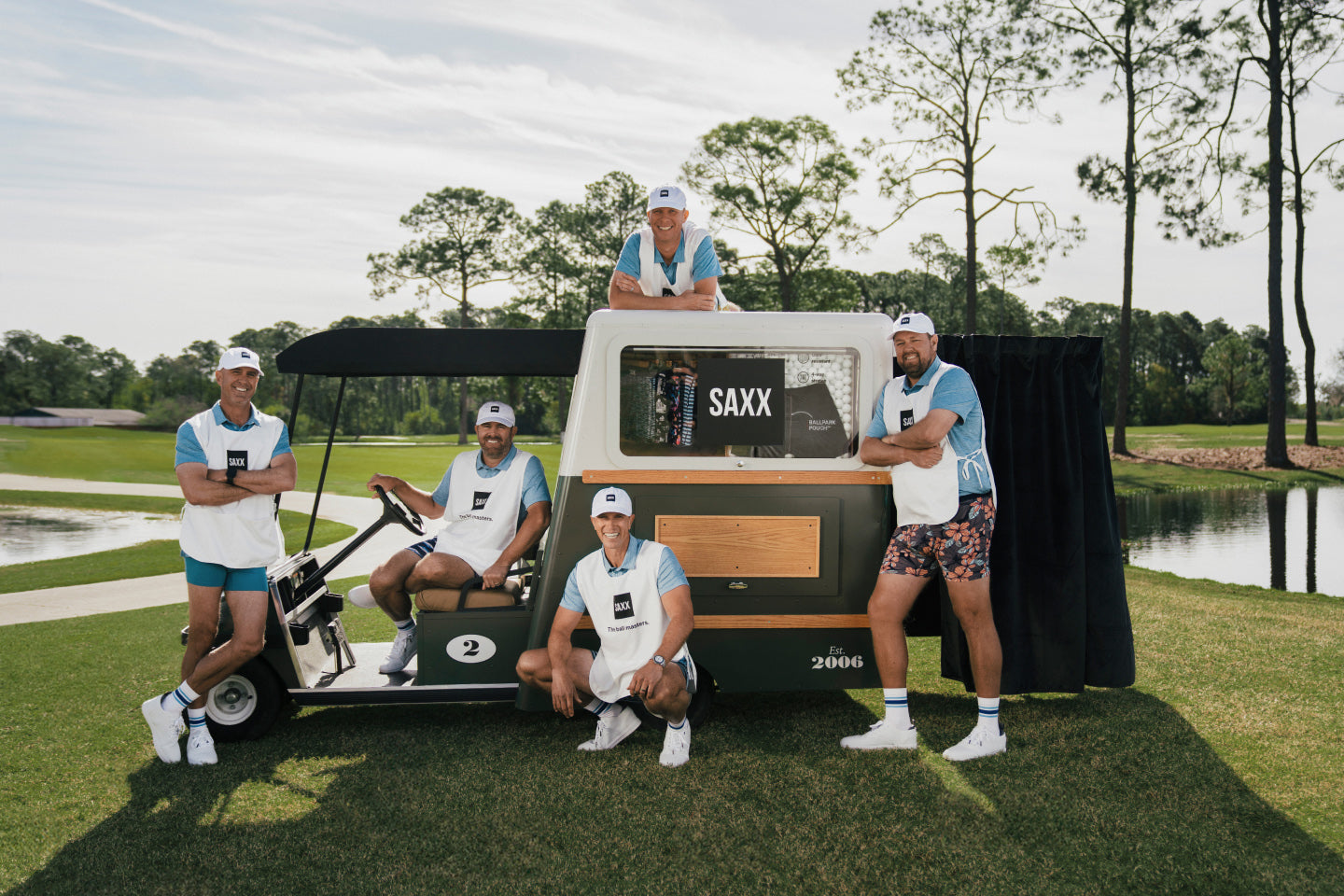Five golf caddies with a golf cart on the fairway. One man sits in the driver's seat while three men flank the cart, and one man lays on top. 