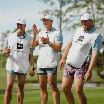 Three men stand on a putting green wearing boxer briefs, blue polos, and white vests. The man in the center is clapping.
