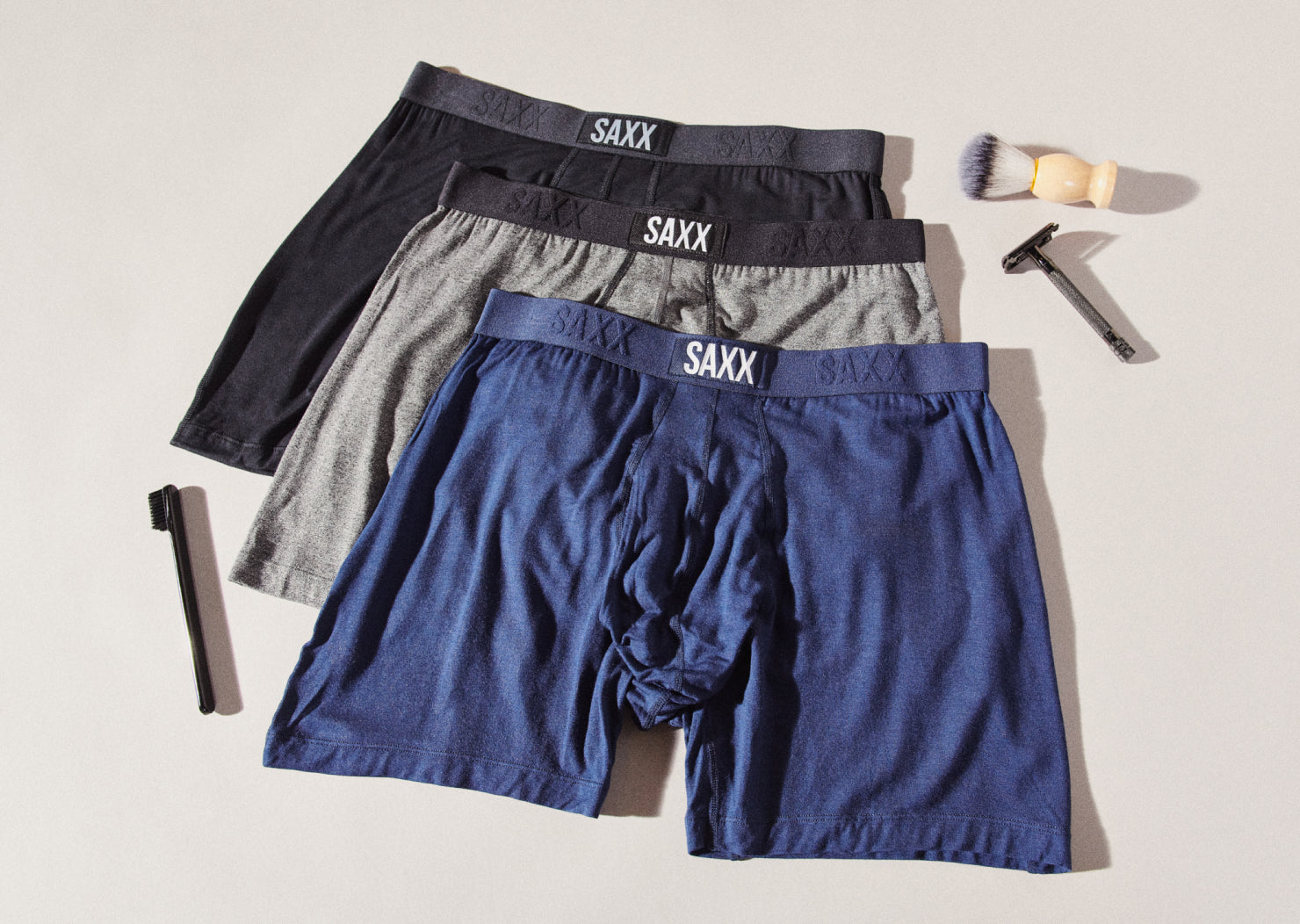 Three pairs of men’s underwear laying on a flat surface. A men’s razor and shaving brush lay on one side of the image. 