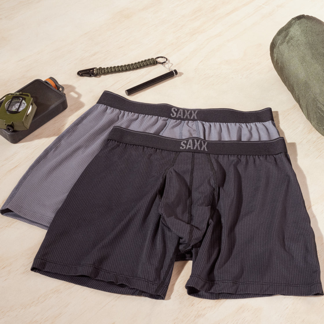 Two pairs of boxer briefs laid on a flat surface. To one side there is a compass on a water cannister and a pen light above. 