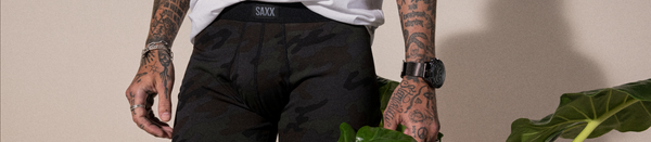 Tattooed man in black camo underwear standing by plant