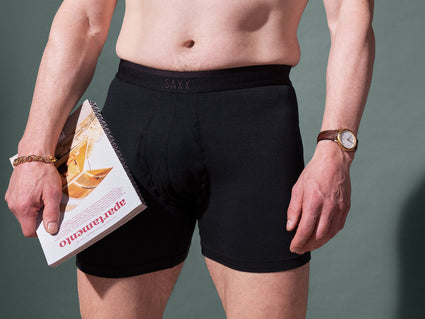 Close-up of a person wearing black SAXX Underwear boxer briefs, holding a book