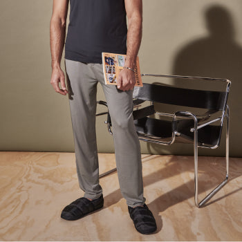 Man in gray silk pants holding a magazine and standing in front of a chair