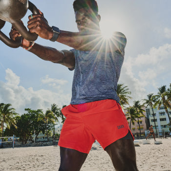 Man in grey shirt and red shorts on beach swinging kettle bells