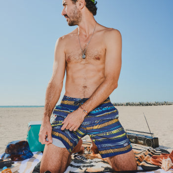 Shirtless man in striped swim shorts kneeling on beach with blanket and stereo