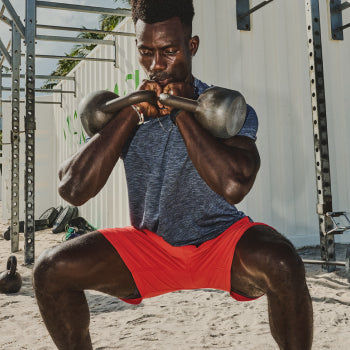 Man in grey shirt and red shorts front squatting two kettlebells on beach