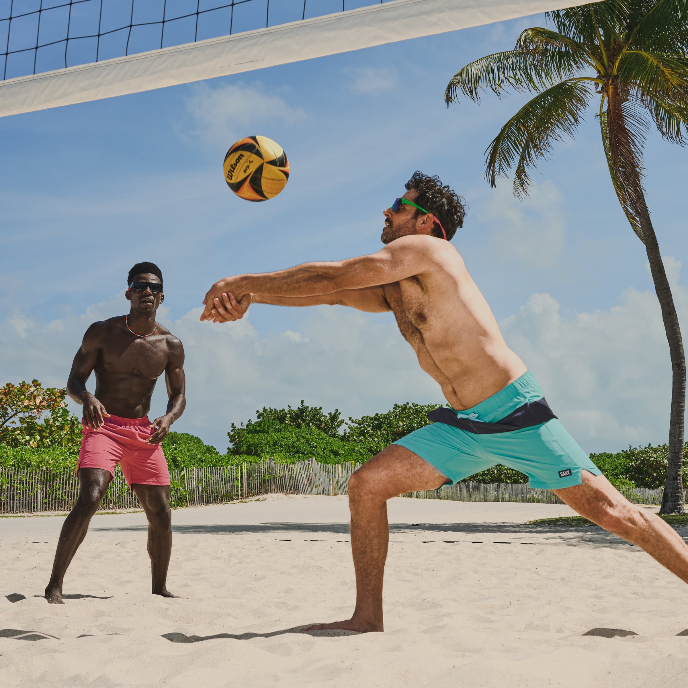 Two men play volley ball on the same side of the court. Both wear bright color swim shorts. The man in the foreground bounces the ball into the air.