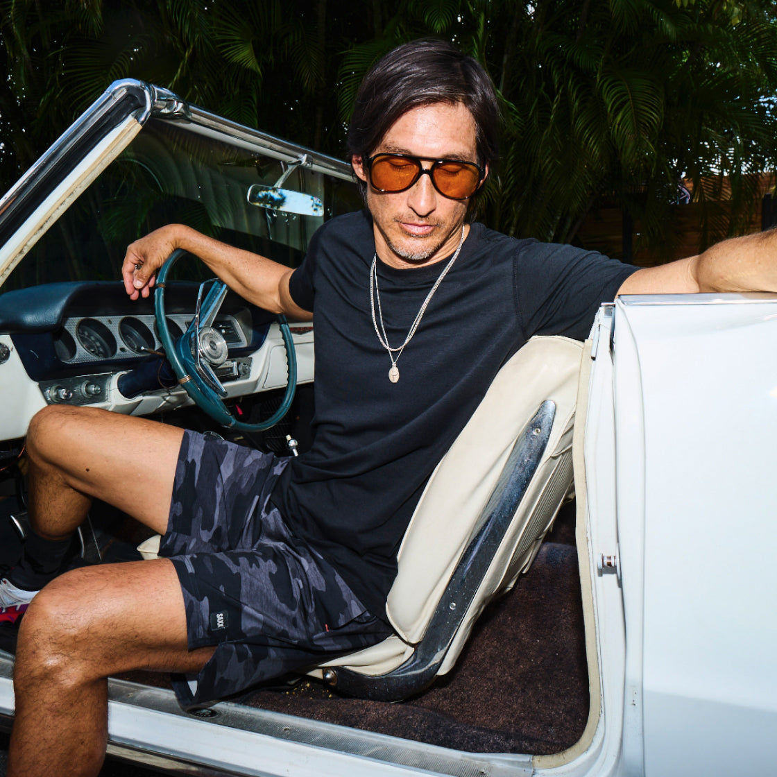 A man with sunglasses on getting out of a vintage sports car wearing athletic shorts and a plain t-shirt in a tropical setting.