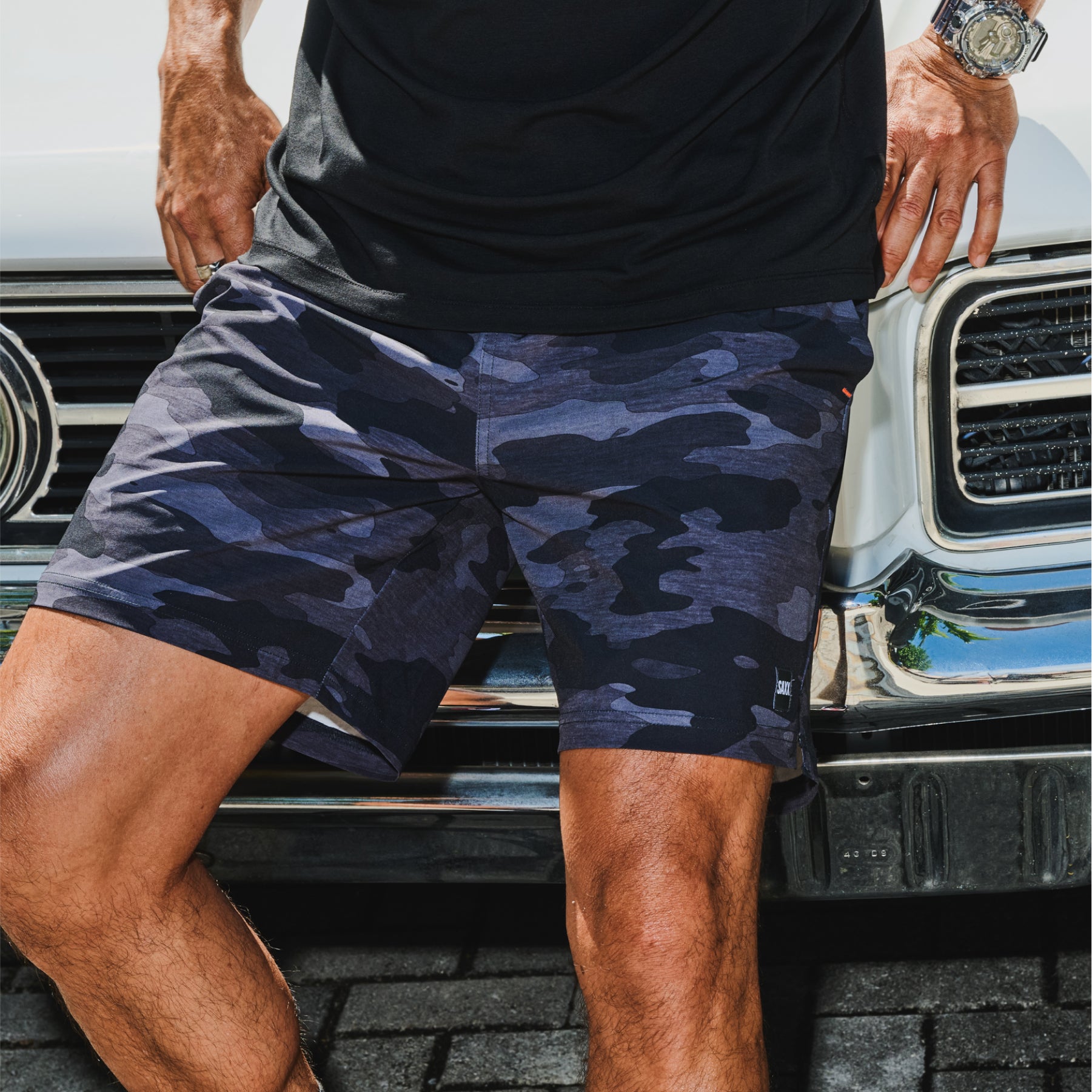 A close up short of a man's camouflage-print sport shorts. The man is leaning against the hood of a classic sports car.