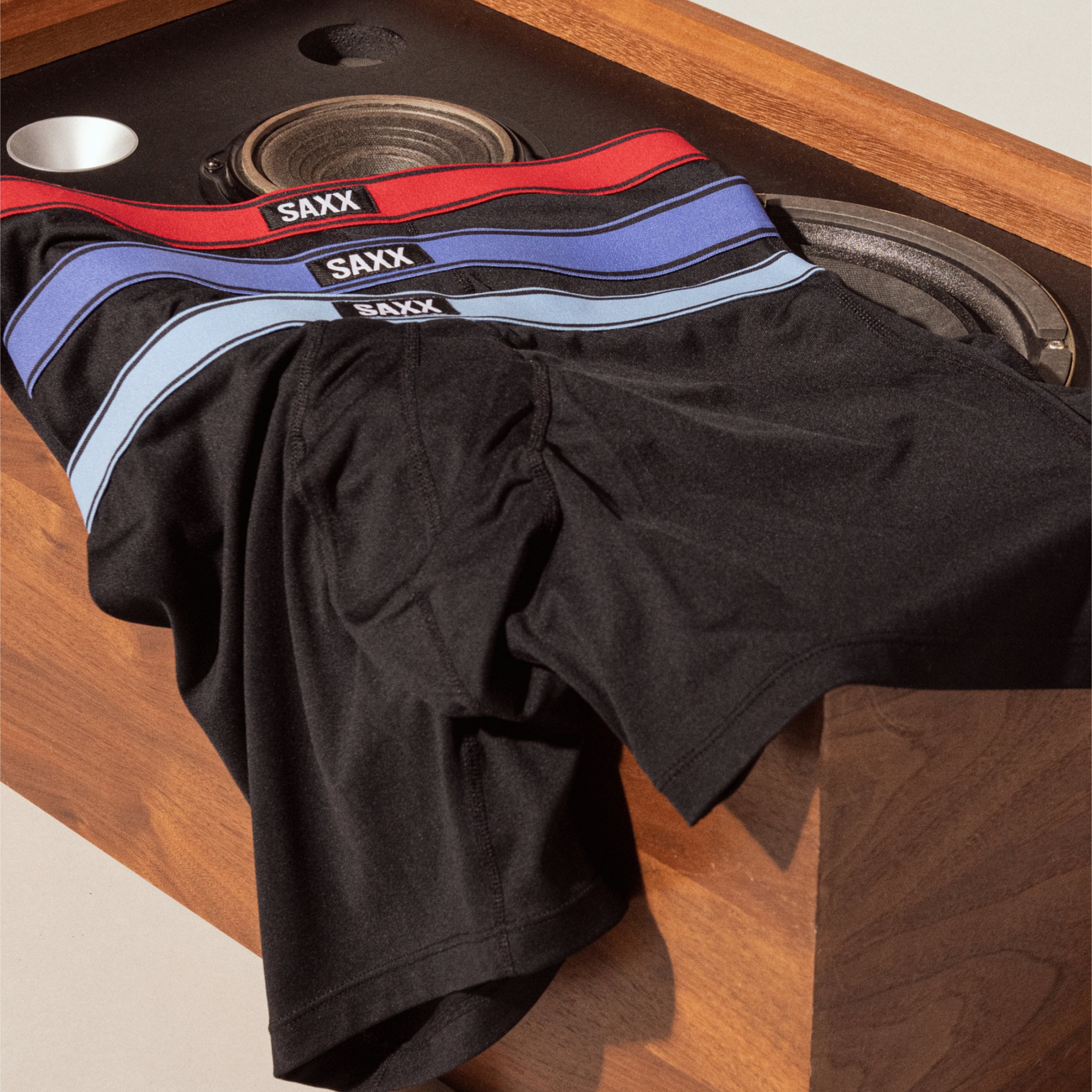 Three pairs of black boxer briefs are laid on a flat surface. Each has a different color waistband: one red, one dark blue, one light blue.