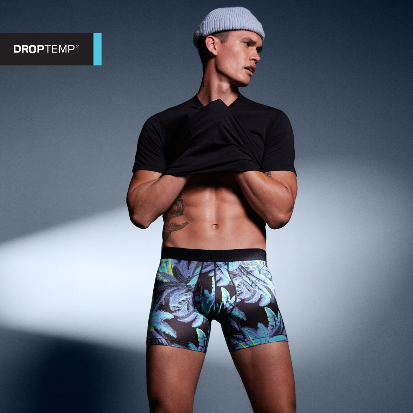 A fit man standing against a neutral backdrop wearing a necklace, black baseball cap, and blue camo-pattern SAXX boxer briefs.  