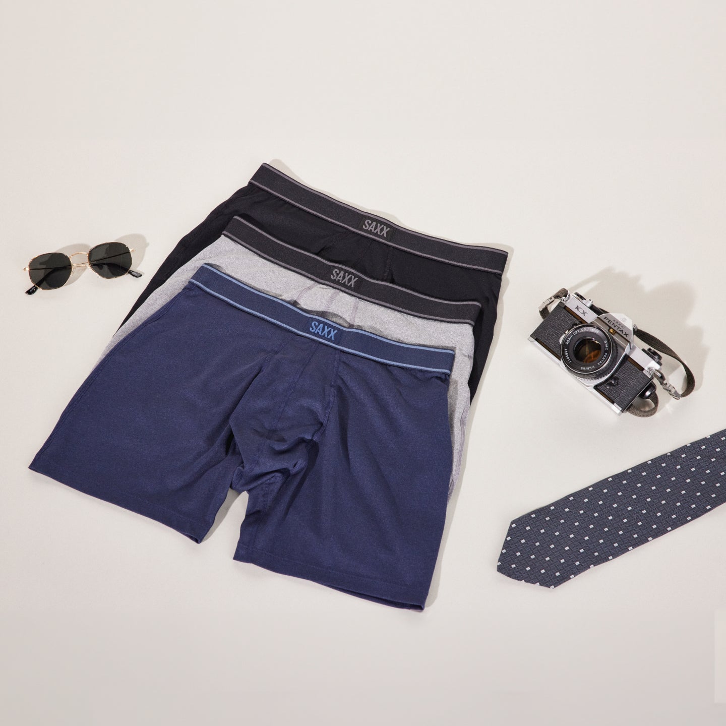 Three pairs of SAXX boxer briefs laid out flat on a white background, surrounded by a pair of sunglasses, a Pentax camera, and a polka-dot tie.