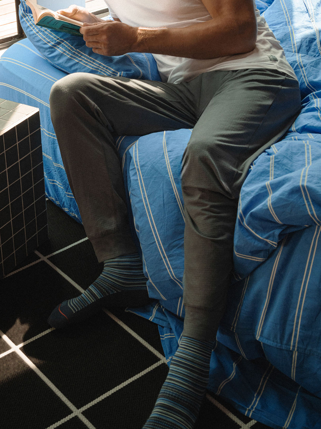 Person sitting on a bed with blue striped bedding, wearing gray pants and socks.