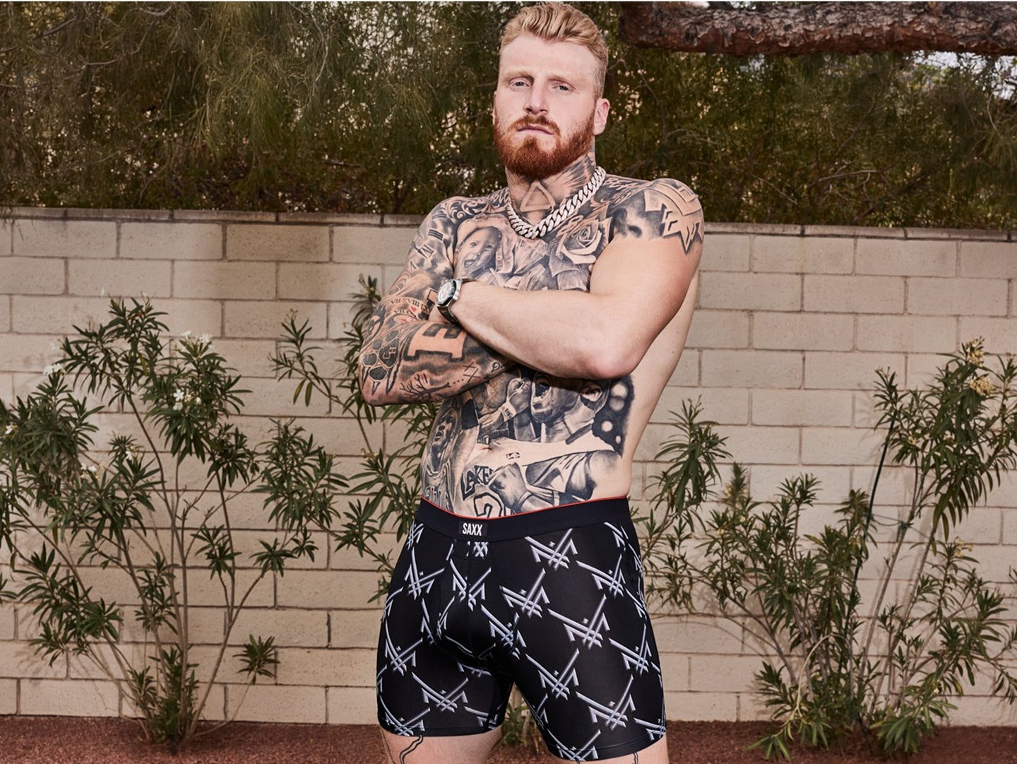 Pro football player Maxx Crosby wearing black patterned shorts standing outdoors against a brick wall.