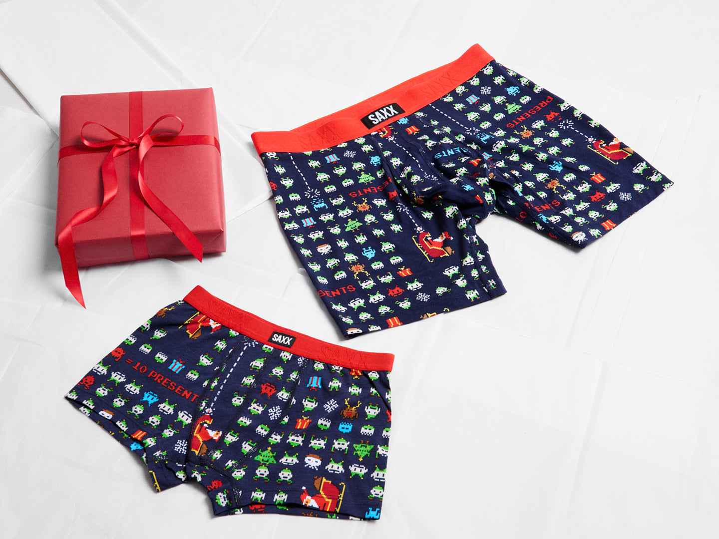 Matching father and son SAXX underwear sets in navy with festive pixel-print pattern and red waistbands, displayed beside red gift boxes - the perfect holiday gift.