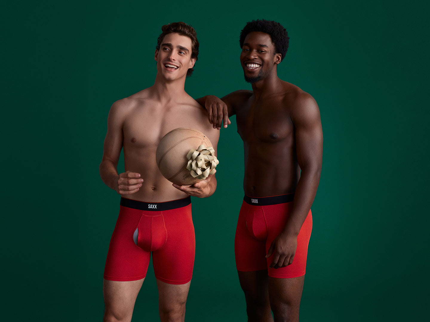 Two men posing in underwear against a green backdrop, while one holds a basketball with a gift bow on it