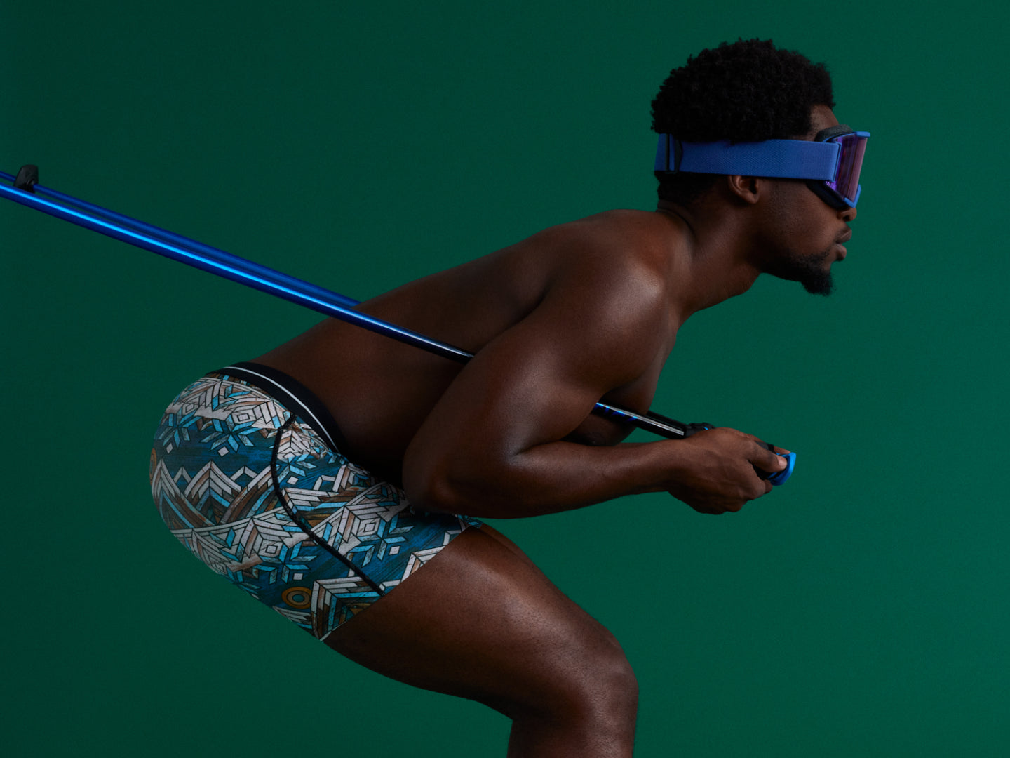 Man wearing patterned swim shorts and goggles, holding a blue pole against a dark green background