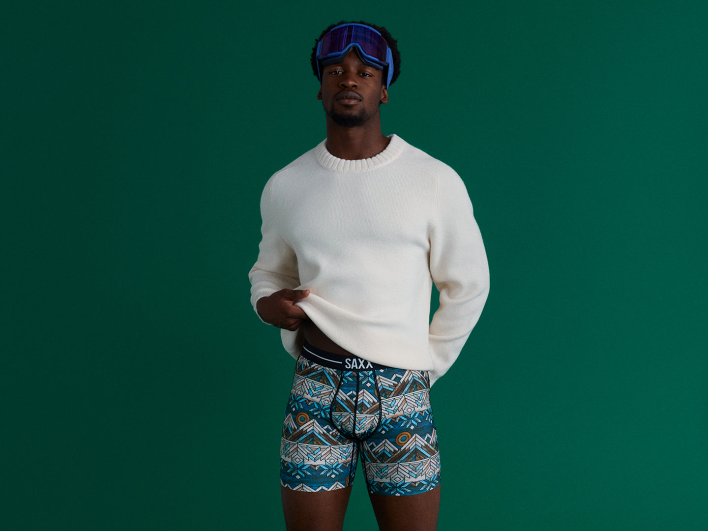 A man in patterned holiday boxer briefs, a white knit sweater, and blue ski goggles posing against a green studio backdrop.