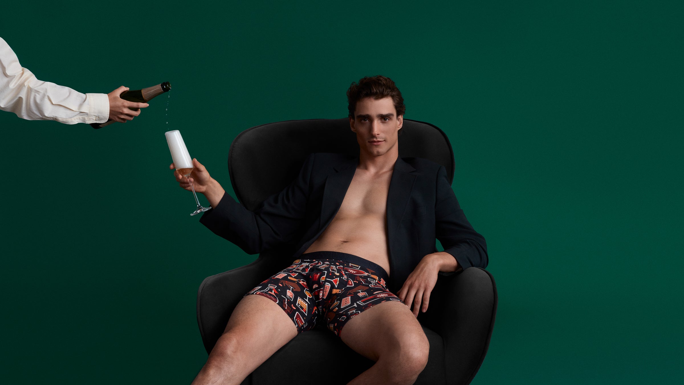 Man in patterned underwear sitting in a chair with a bottle and glass held by another person against a green background