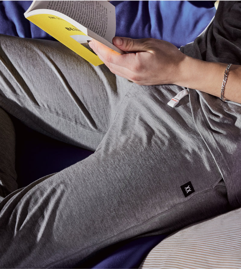 Person wearing gray pajama pants with a small logo, sitting and reading a book.