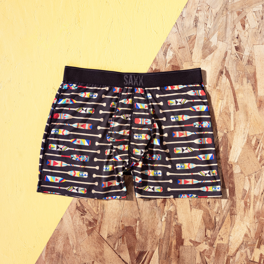 Black SAXX Boxer Briefs with horizontal multi-color geometric-pattern canoe paddles, laid out flat over pallet wood surface.