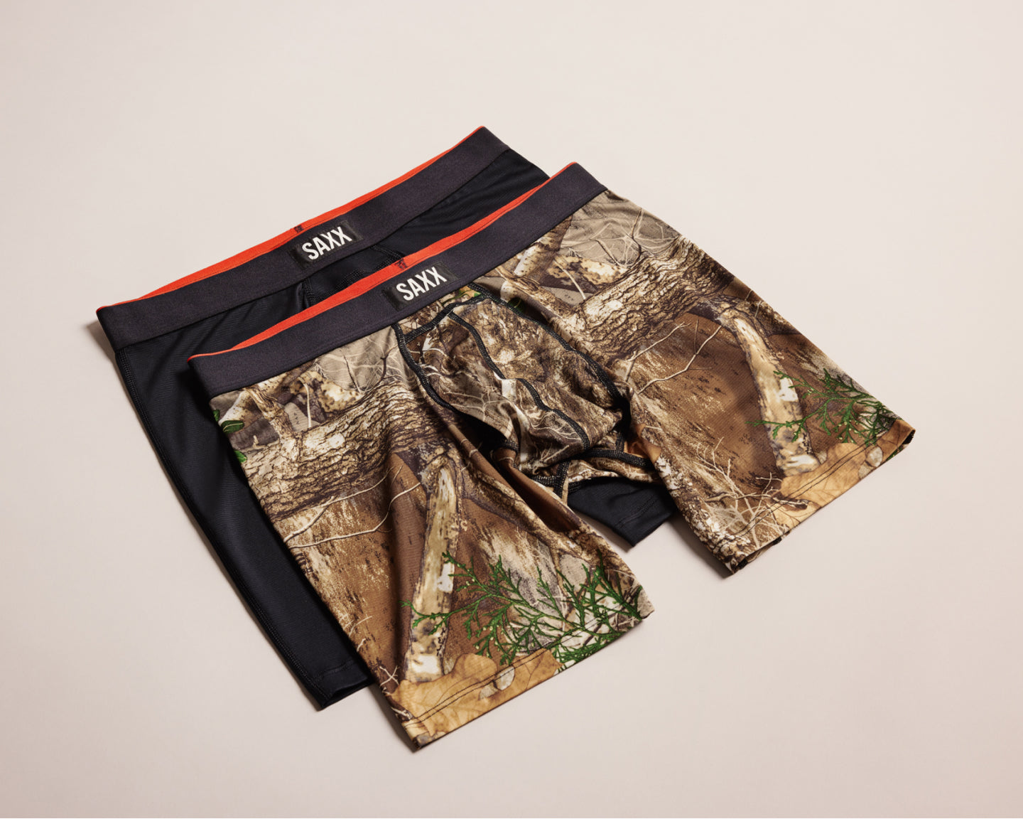 Two pairs of SAXX underwear, one black and one camouflage patterned, on a beige background.