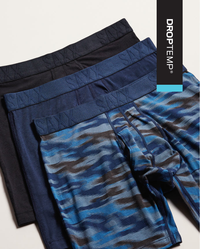 Three pairs of men's boxer briefs from DROPTEMP are stacked diagonally. The front pair features a blue, gray, and brown abstract camouflage pattern, while the back pairs are solid navy and black.