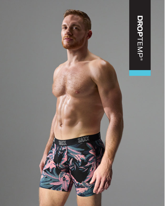 A male model wearing DROPTEMP brand boxer briefs with a pink and teal tropical leaf pattern on a black background, posed against a solid gray studio backdrop.
