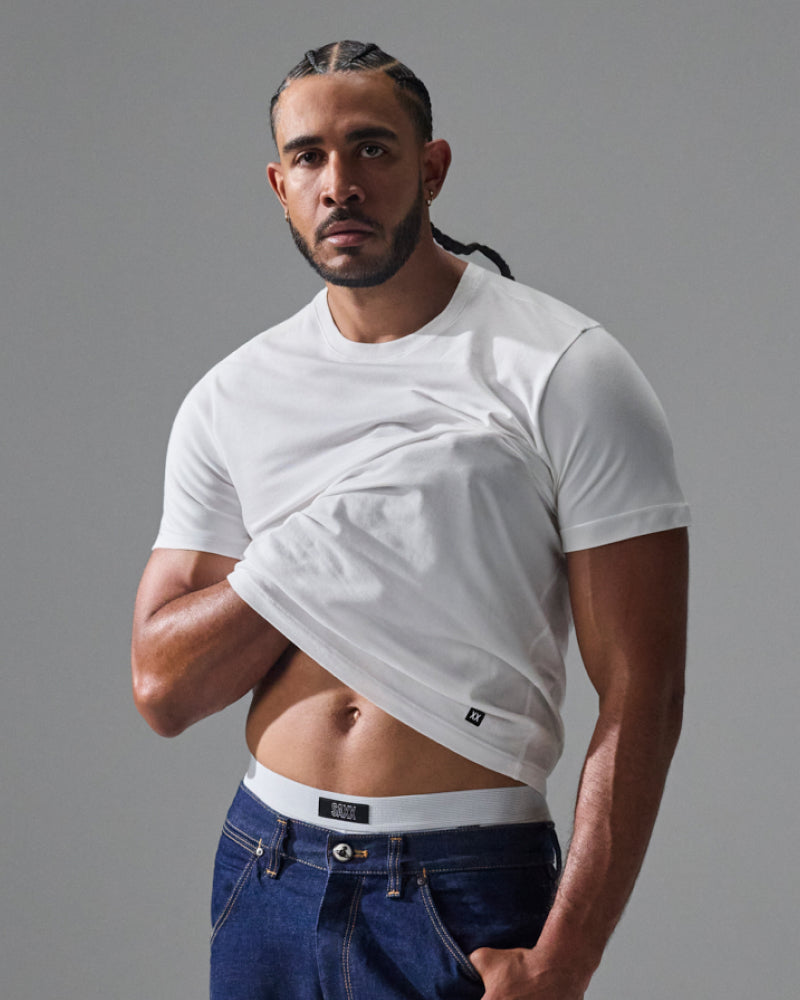 A muscular man with braided hair wearing a white short-sleeve crewneck t-shirt and blue jeans, lifting the shirt slightly to show the fit and drawstring-free waistband, against a solid gray studio background.