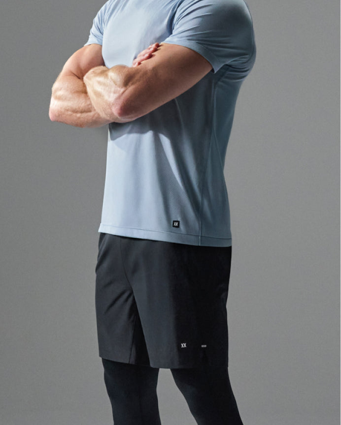 A man wearing a light blue athletic t-shirt and black athletic shorts, shown from the chest down against a neutral gray studio background.