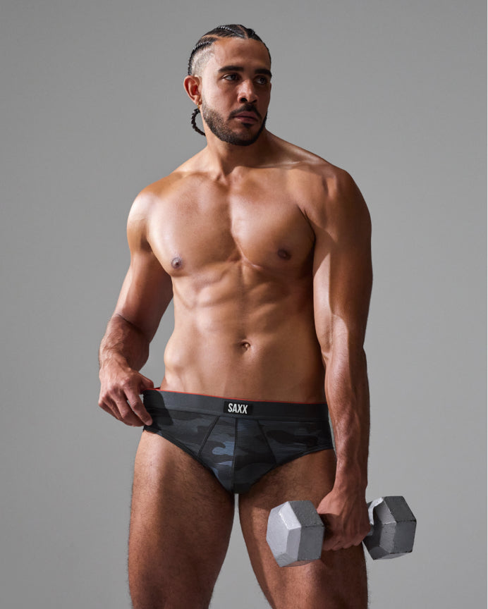 A shirtless male model stands against a gray studio background wearing dark camouflage briefs with a black waistband and the brand name 'SAXY' printed in white. He is holding a gray hexagonal dumbbell.