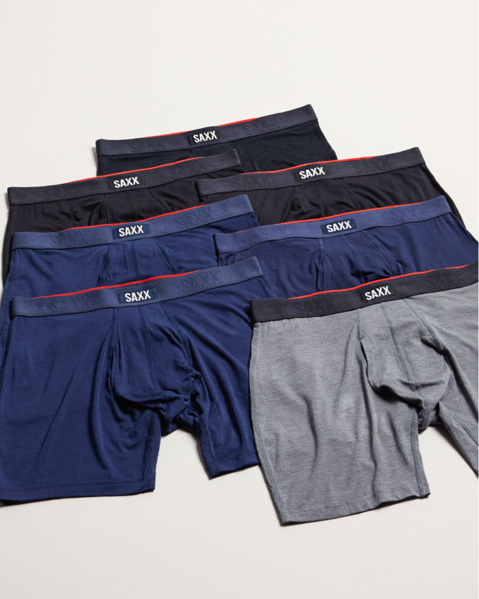 Six pairs of SAXX men's boxer briefs in navy, black, and heather gray, arranged in a fan-like stack on a light beige background.