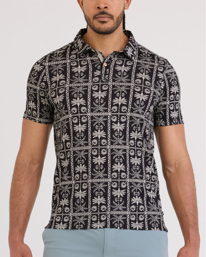 A man wearing a short-sleeve polo shirt with a black and white geometric block print pattern, paired with light blue pants, against a plain light gray background.