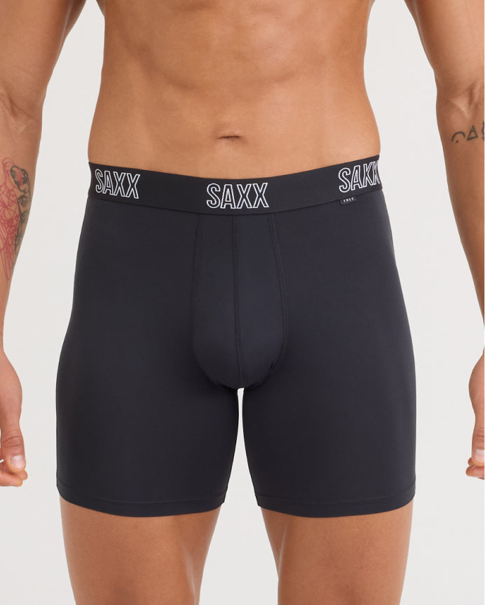 A close-up of a male model's lower torso wearing SAXX brand boxer briefs in black. The underwear has a wide black waistband with the white 'SAXX' logo printed three times, and is photographed against a clean, light gray background.