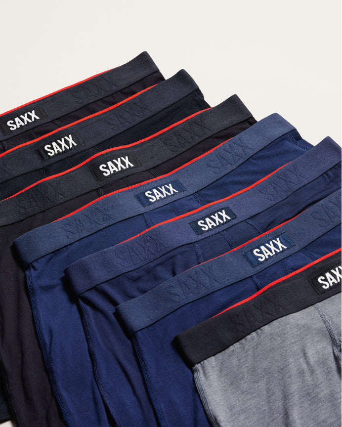 A close-up, angled view of several folded pairs of SAXX men's boxer briefs in navy, blue, black, and gray, showing their elastic waistbands with the brand name 'SAXX' in white.