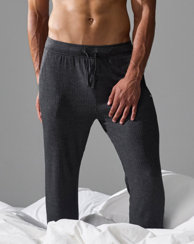 A man wearing dark charcoal gray ribbed knit lounge pants, standing on white rumpled bedding against a solid gray background.