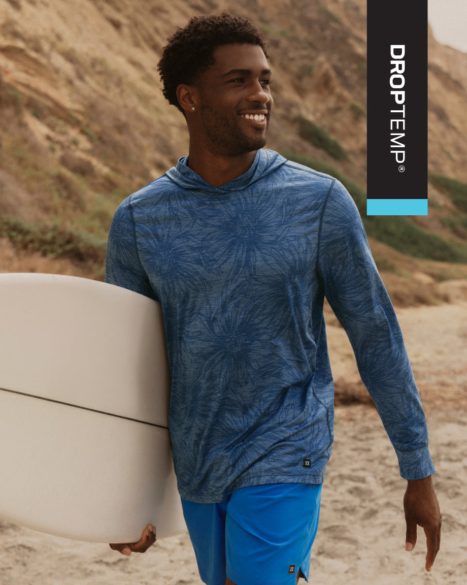Man wearing a blue hoodie and shorts holding a surfboard on a beach with 'Droptemp' branding.