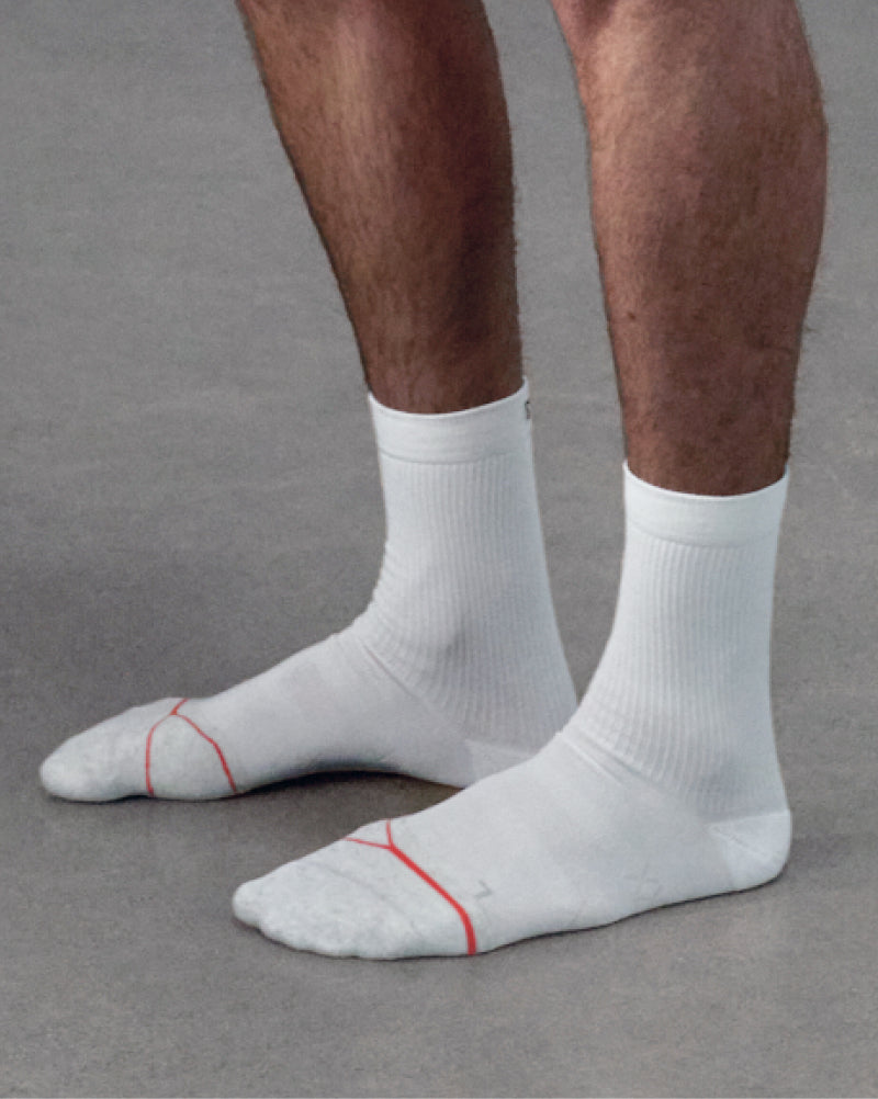 A close-up of a person's feet and lower legs wearing white athletic crew socks with thin red accent lines on the toes, standing on a gray floor.