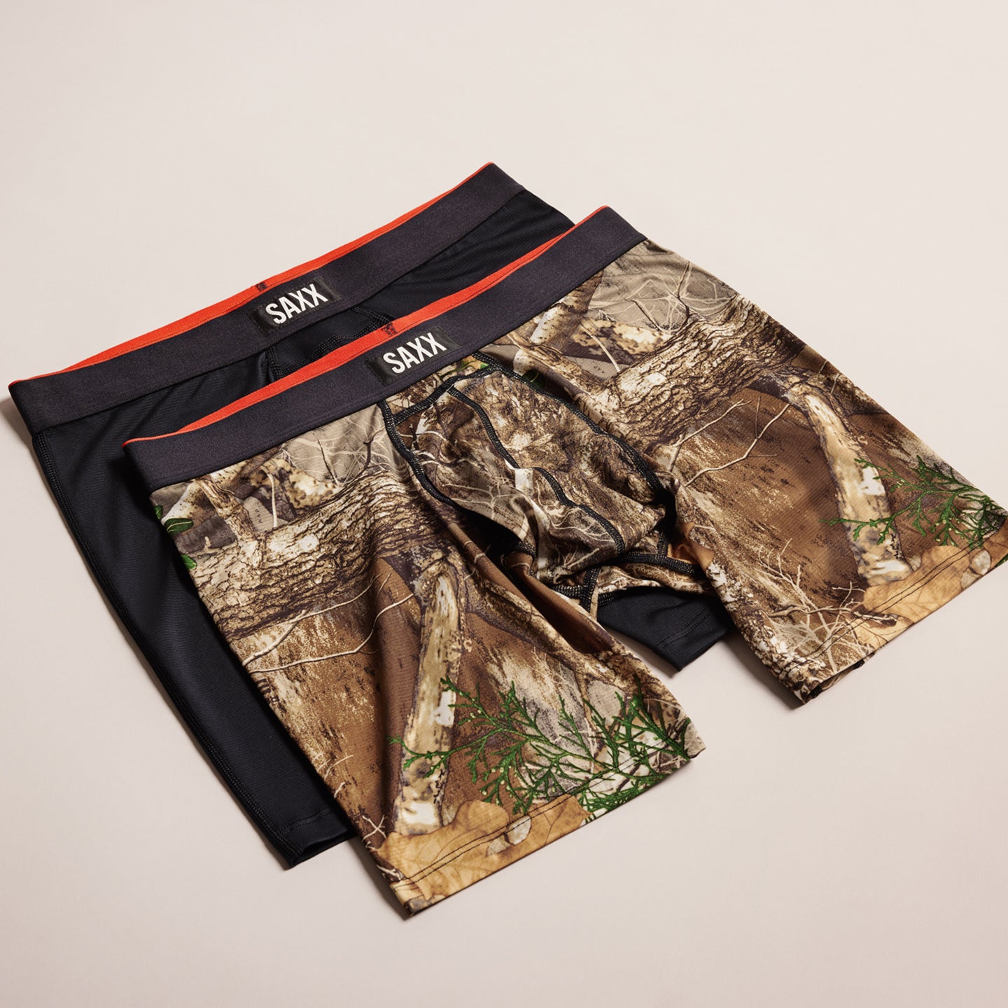 Two pairs of SAXX underwear, one black and one camouflage patterned, on a beige background.