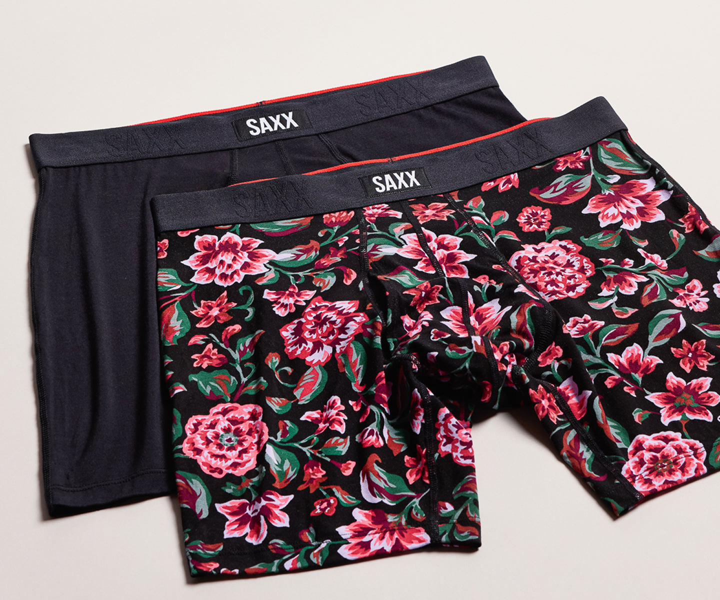 Three pairs of SAXX men's boxer briefs arranged in an overlapping flat-lay. The front pair features a vibrant pink and red floral print on a black background, the middle pair is solid black with a thin red waistband, and the back pair is solid black with a thin red waistband.