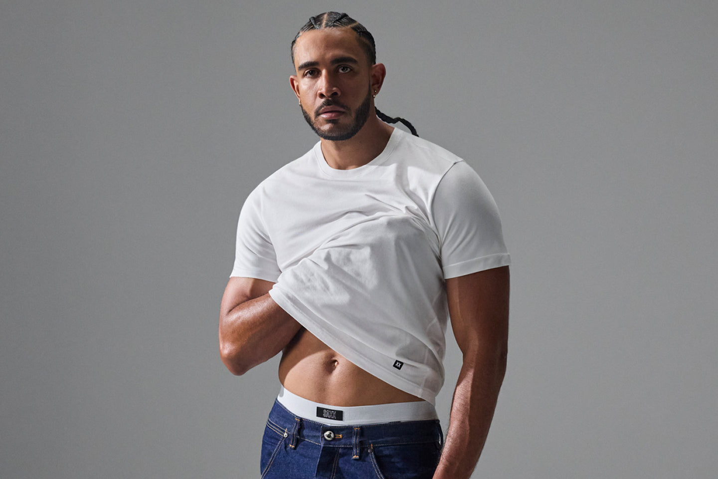 A muscular man with braided hair wearing a white cropped t-shirt and blue jeans, posing against a solid gray studio background.