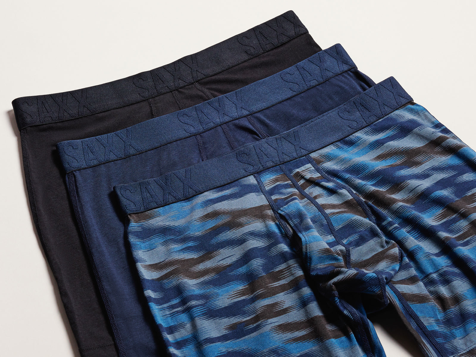 Three pairs of men's boxer briefs stacked diagonally on a light beige background. The front pair features a blue, gray, and brown abstract wavy pattern, the middle pair is solid navy blue, and the back pair is solid black.