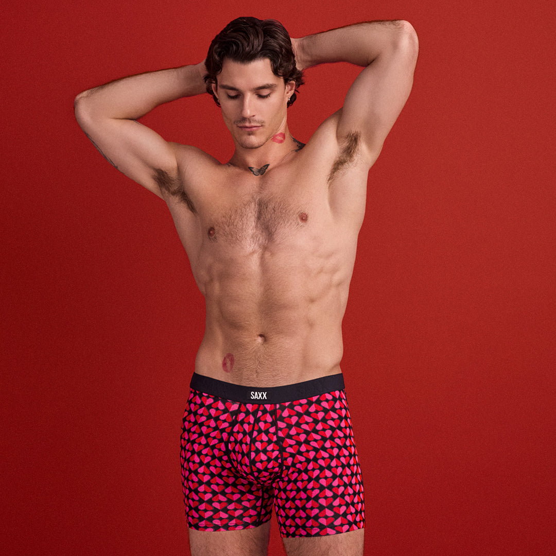 Man wearing pink geometric patterned boxer briefs with 'SAXX' brand logo on a red background