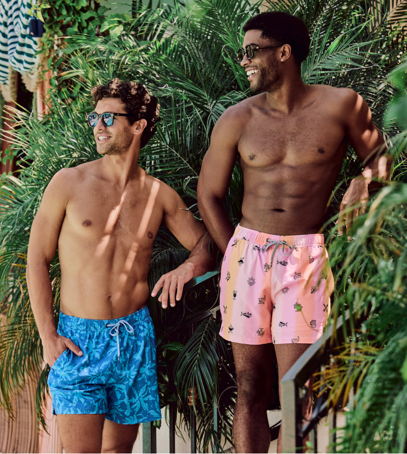 Two men standing in swim trunks in front of large green plant