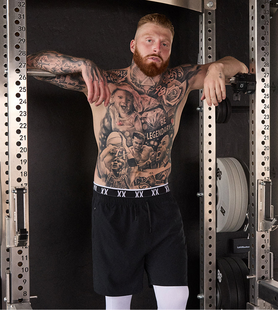 Pro Football Player Maxx Crosby stands inside a power rack wearing black athletic shorts with a white elastic waistband featuring the brand name 'XX' and white compression sleeves. The background is a dark gym setting with weight plates on the rack.