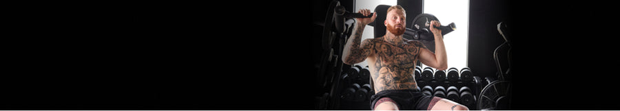 Pro football player Maxx Crosby in a gym setting holding a barbell overhead, with a bright light background and dark gym equipment around him.