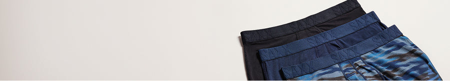 Three pairs of men's boxer briefs stacked on a light beige background, showing waistbands and fabric textures in black, navy, and blue with a subtle pattern.