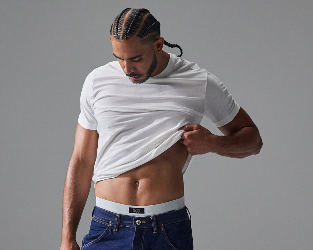Man wearing a white SAXX undershirt and blue jeans against a solid background